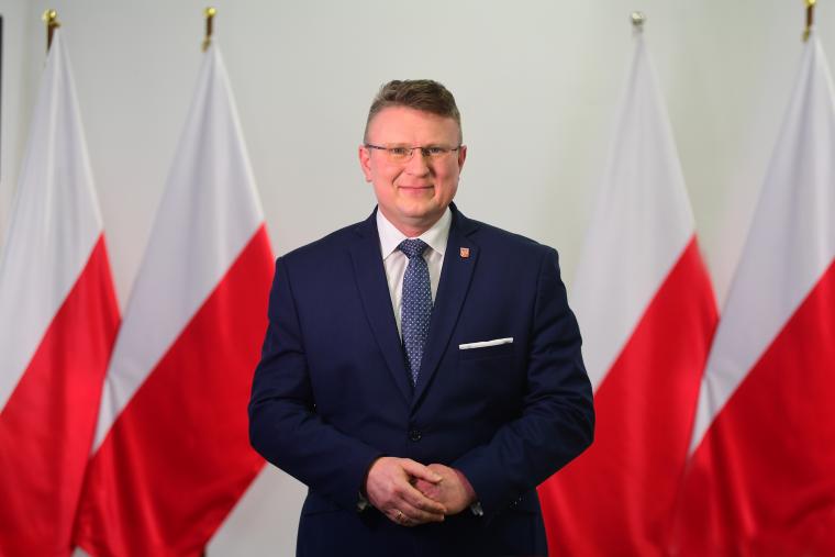 Arkadiusz Kubich - zdjęcie przedstawia mężczyne w garniturze na tle flag Polski